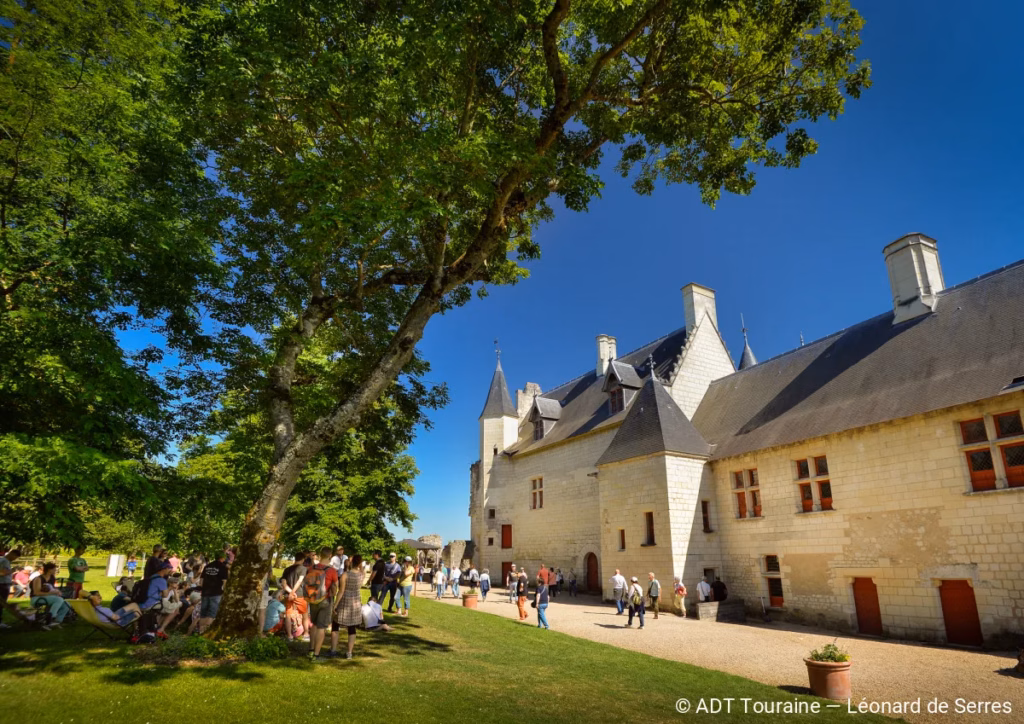 Stone château with steep slate roofs and turrets set in a sunny courtyard, groups of visitors gathered on the lawn under a large leafy tree.
