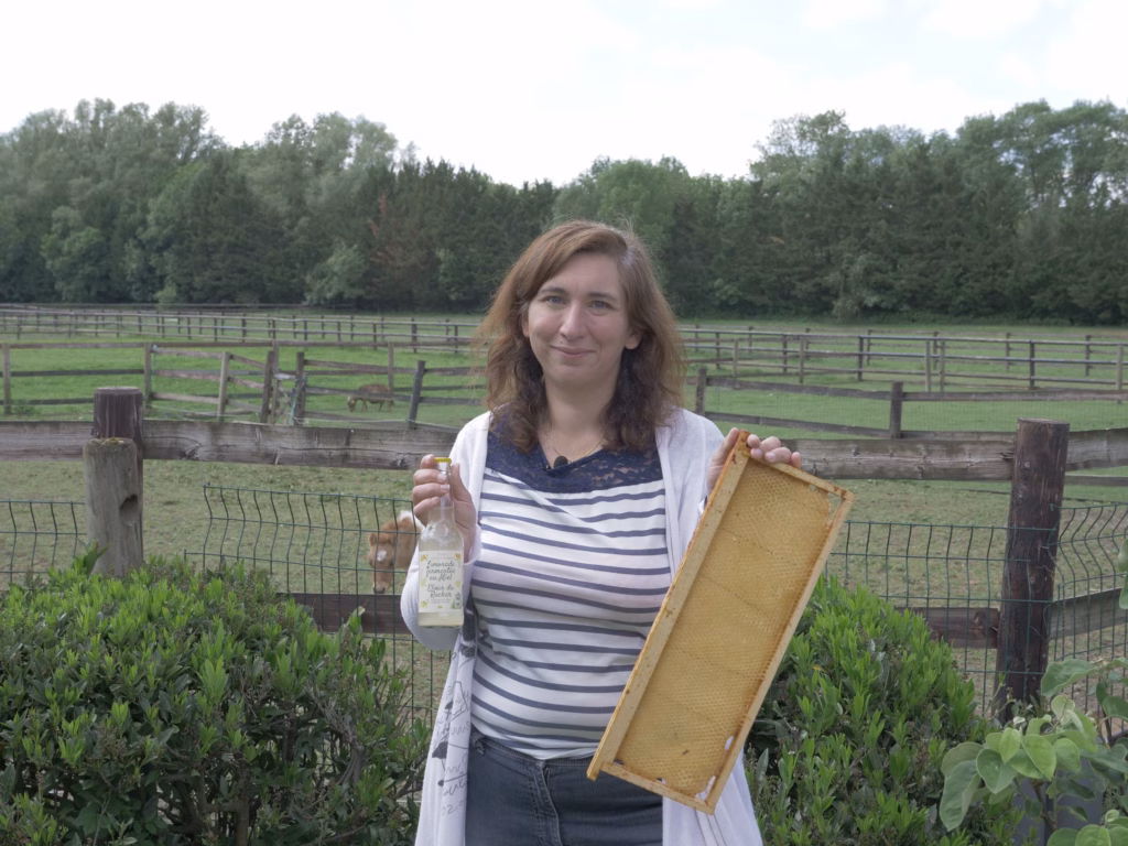 Smiling woman in a striped shirt stands by a wooden fence in a farm field, holding a honeycomb frame and a bottle of honey with green hedges in the foreground.