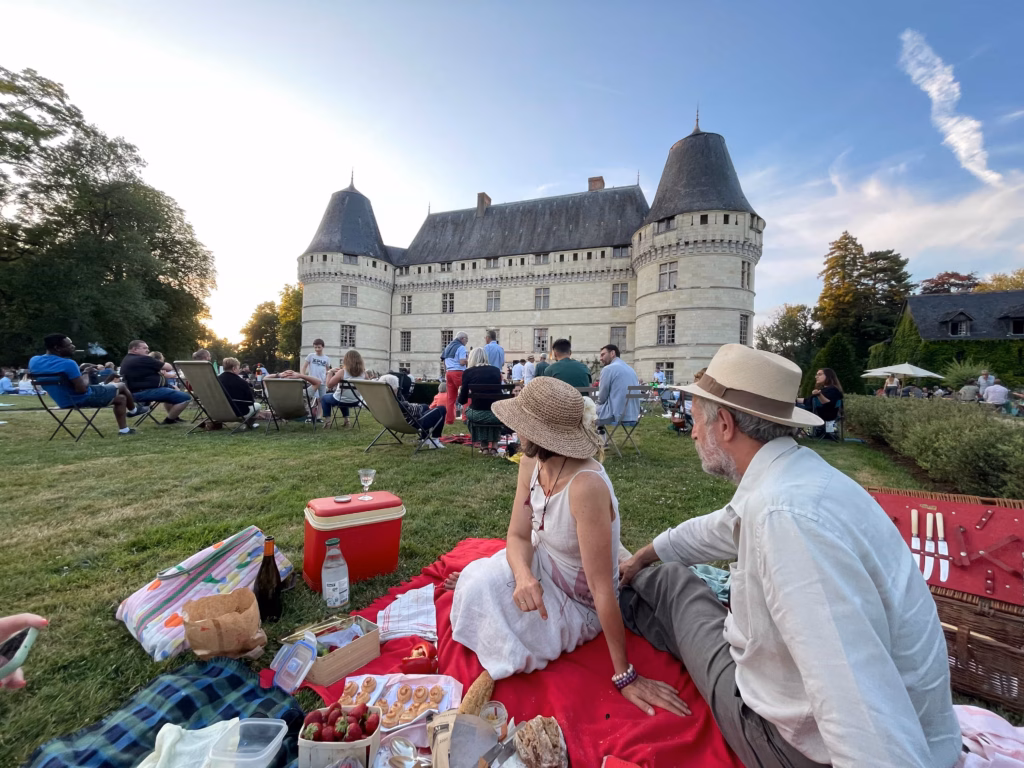 People picnicking on a grassy lawn in front of a historic castle with rounded towers and a crowd relaxing on chairs and blankets.