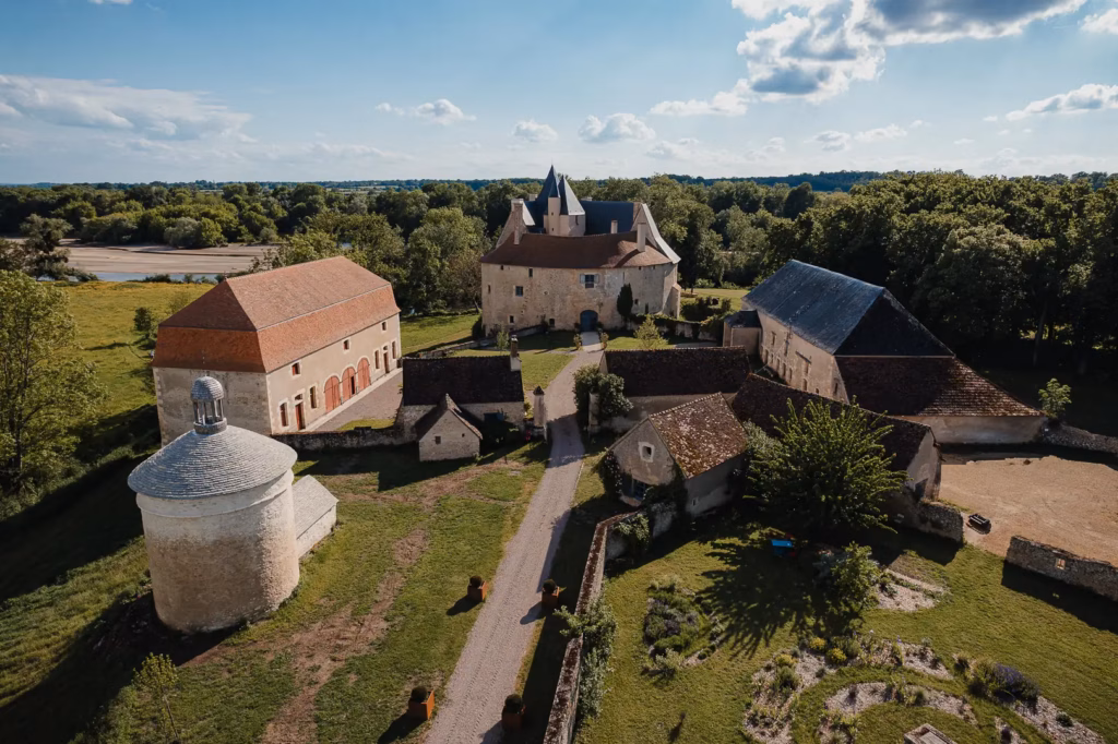 Vue aérienne d’un complexe de château médiéval comprenant des bâtiments en pierre, une tour ronde et des toits en tuiles rouges, entourés d’arbres verdoyants, sous un ciel bleu.