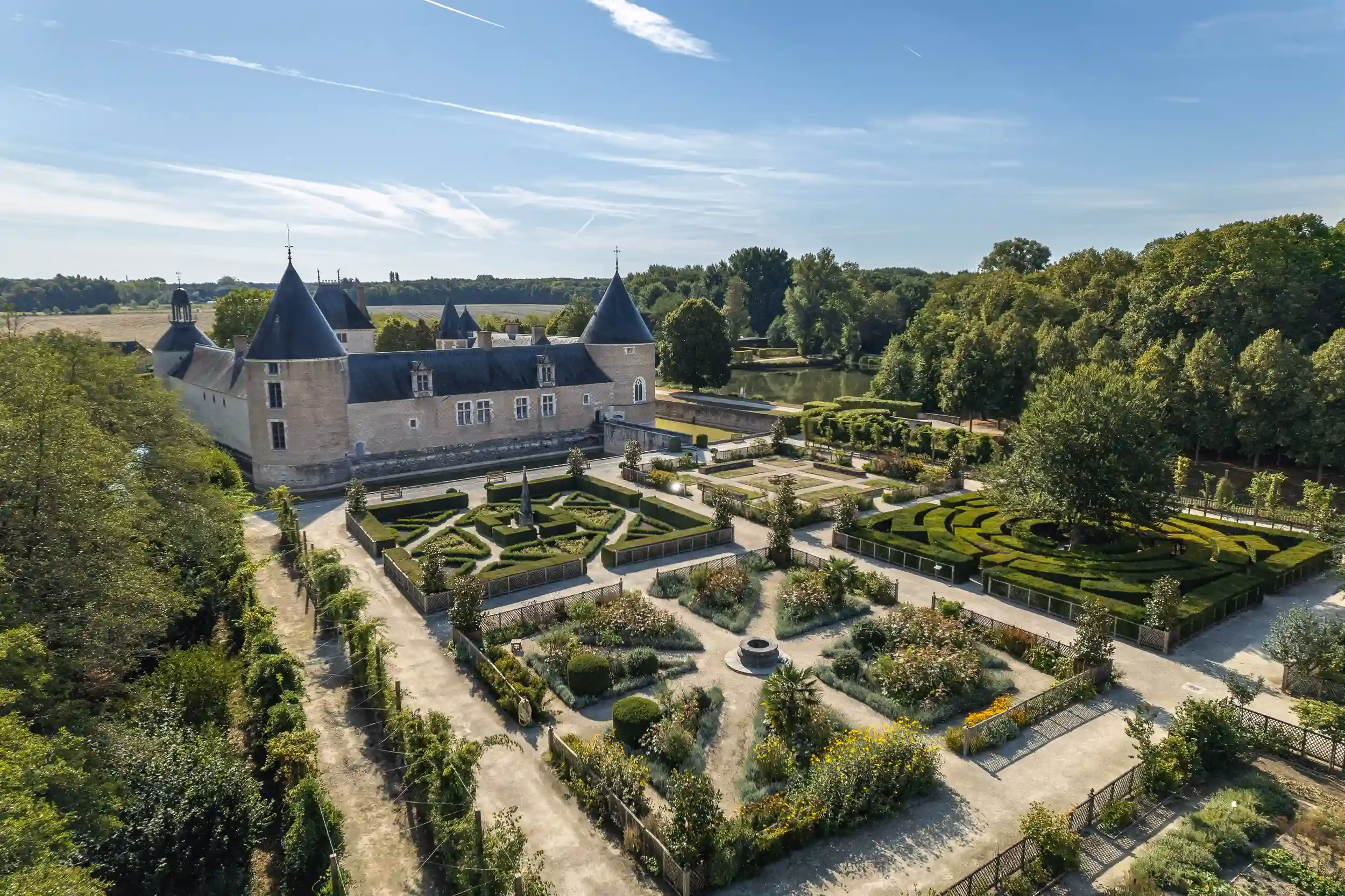 A historic stone castle with conical towers and formal manicured gardens viewed from above, under a blue sky.