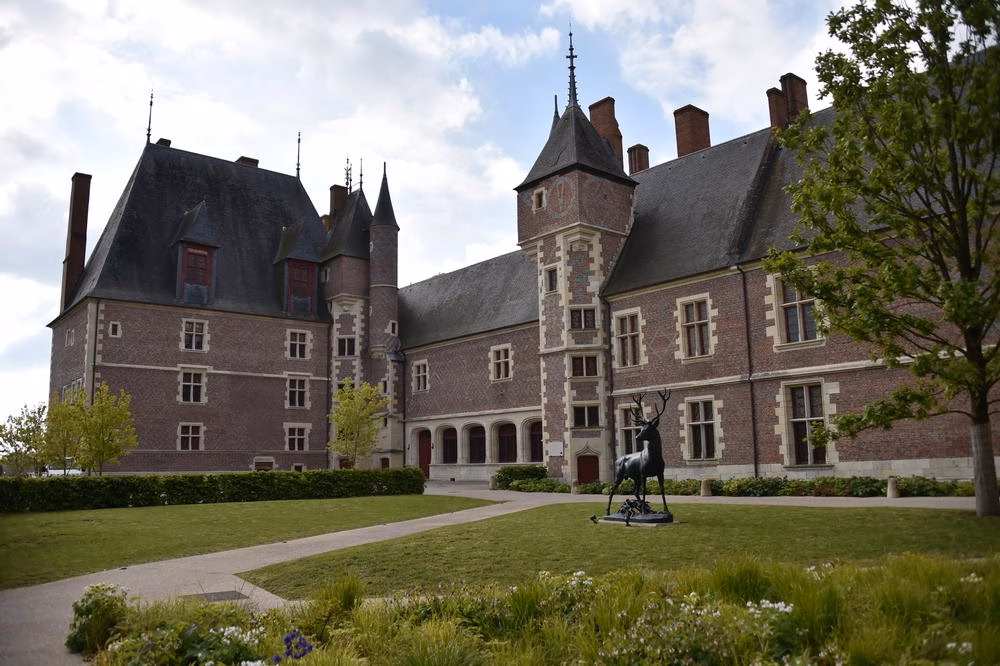 Brick castle with towers and slate roofs, a manicured lawn, and a black stag statue in the foreground.